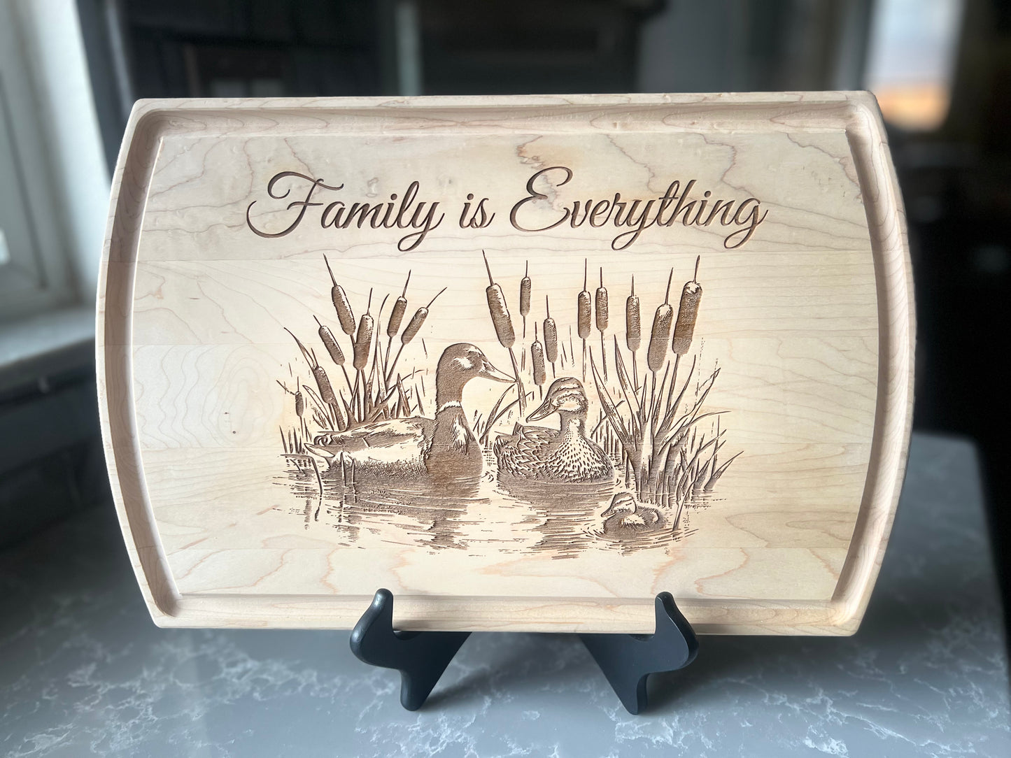 Maple wooden cutting board with engraved ducks and 'Family is Everything' text on a stand.